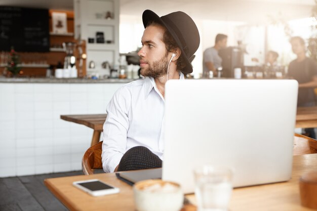 Joven hipster barbudo con sombrero de moda escuchando música o audiolibro con auriculares blancos, sentado en una mesa de madera con una computadora portátil y un teléfono celular con pantalla en blanco durante el almuerzo en la acogedora cafetería