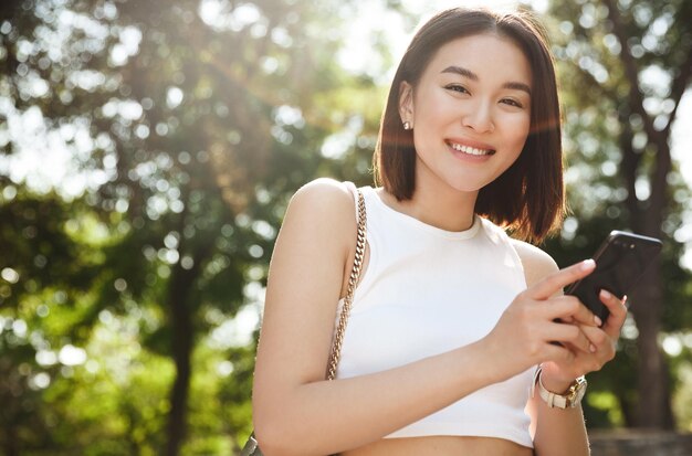 La joven hipster está chateando a través de un teléfono móvil en la red con sus amigos. Una mujer bonita está leyendo un mensaje de texto en su teléfono celular mientras se relaja en el parque.