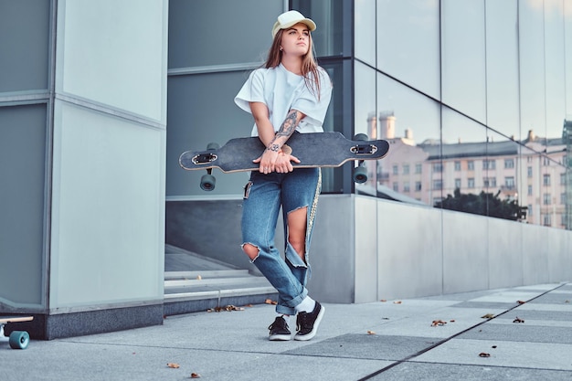 Una joven hipster con gorra vestida con camisa blanca y jeans rasgados sostiene una patineta mientras posa cerca de un rascacielos.