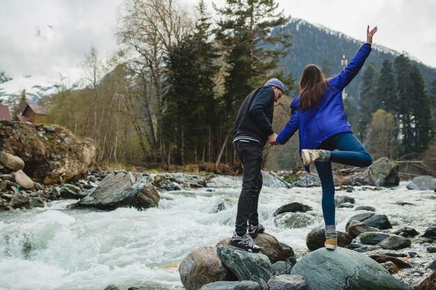 Joven hipster hermosa pareja de enamorados caminando sobre una roca en el río en el bosque de invierno