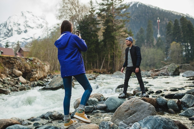 Joven hipster hermosa pareja de enamorados sosteniendo smartphone, tomando fotografías, en el río en el bosque de invierno