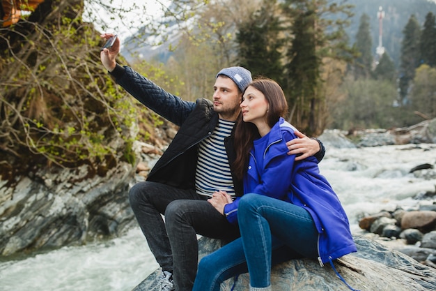 Joven hipster hermosa pareja de enamorados sosteniendo smartphone, tomando fotografías, sentado sobre una roca en el río en el bosque de invierno