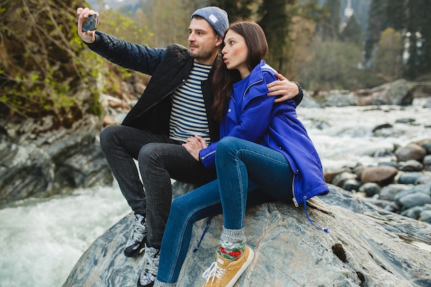 Joven hipster hermosa pareja de enamorados sosteniendo smartphone, tomando fotografías, sentado sobre una roca en el río en el bosque de invierno