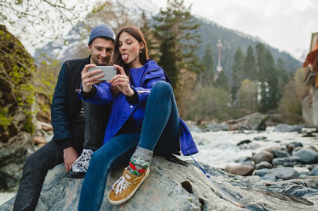 Joven hipster hermosa pareja de enamorados sosteniendo smartphone, tomando fotografías, sentado sobre una roca en el río en el bosque de invierno