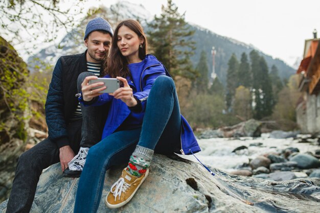 Joven hipster hermosa pareja de enamorados sosteniendo smartphone, tomando fotografías, sentado sobre una roca en el río en el bosque de invierno