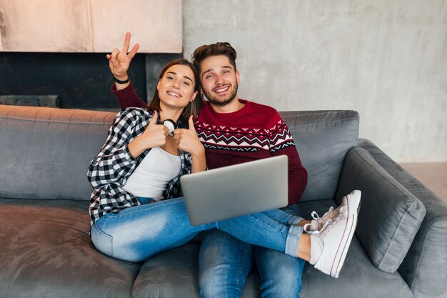 Joven hipster hombre y mujer sonrientes sentados en casa en invierno, sosteniendo el portátil