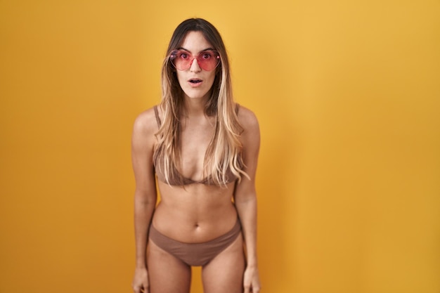 Joven hispana con bikini sobre fondo amarillo asustada y conmocionada con sorpresa y expresión asombrada miedo y cara emocionada