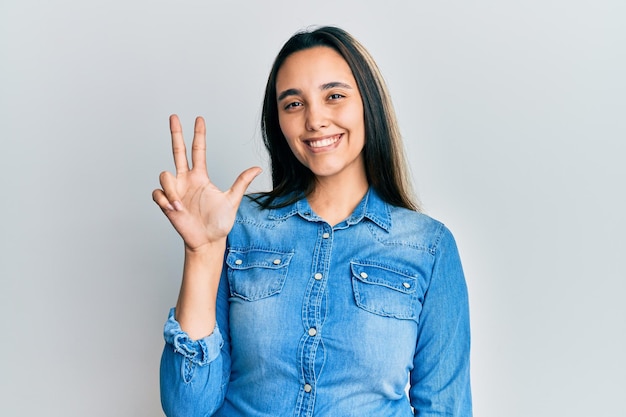 Joven hispana con chaqueta de mezclilla casual que aparece y señala con los dedos número tres mientras sonríe confiada y feliz