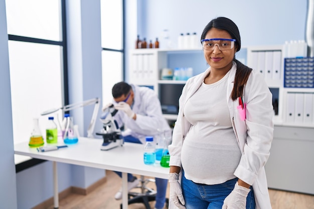 Joven hispana esperando un bebé trabajando en un laboratorio científico con expresión seria en la cara simple y natural mirando a la cámara