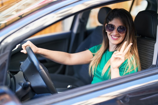 Joven hispana haciendo gesto de victoria conduciendo un coche en la calle