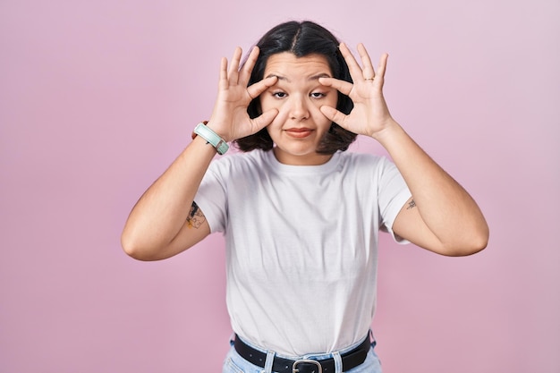 Foto gratuita joven hispana con pantalones blanco casual sobre fondo rosa tratando de abrir los ojos con los dedos, somnolienta y cansada por la fatiga matutina