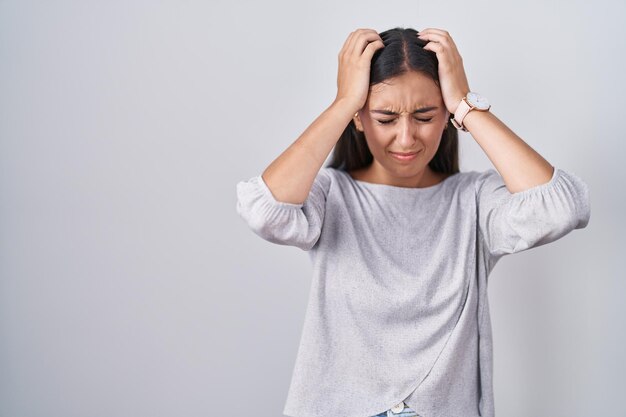 Joven hispana parada sobre fondo blanco que sufre de dolor de cabeza desesperada y estresada por el dolor y la migraña. manos en la cabeza.