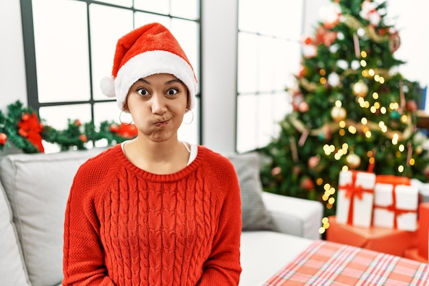 Joven hispana con el pelo corto y sombrero de navidad sentada en el sofá hinchando las mejillas con cara graciosa. boca inflada de aire, expresión loca.