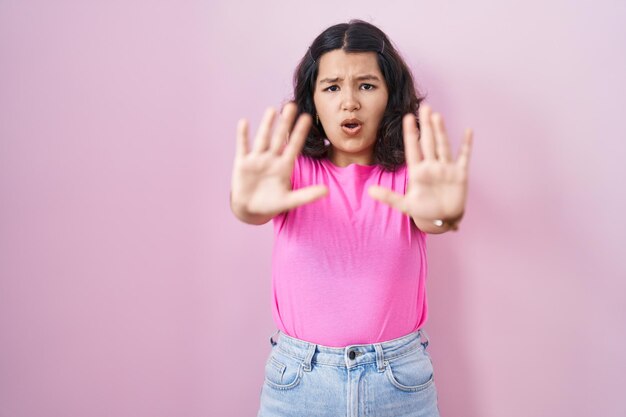 Joven hispana de pie sobre fondo rosa haciendo un gesto de parada con las palmas de las manos, expresión de enojo y frustración