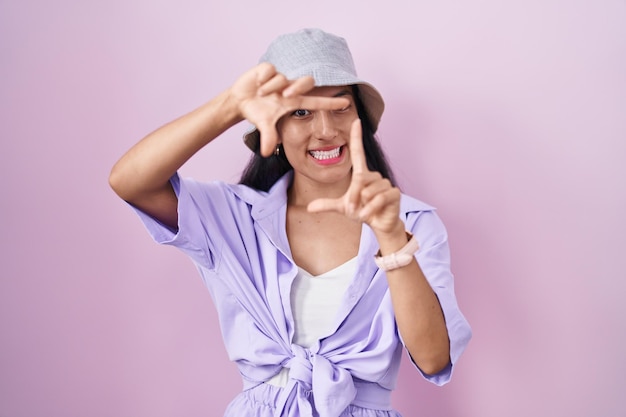 Foto gratuita joven hispana de pie sobre fondo rosa con sombrero sonriendo haciendo marco con manos y dedos con cara feliz creatividad y concepto de fotografía