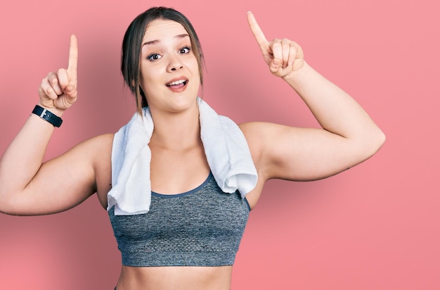 Joven hispana con ropa deportiva y toalla sonriendo asombrada y sorprendida y señalando con los dedos y los brazos levantados.