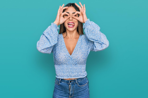 Foto gratuita joven hispana con ropa informal haciendo un buen gesto como binoculares sacando la lengua a los ojos mirando a través de los dedos expresión loca
