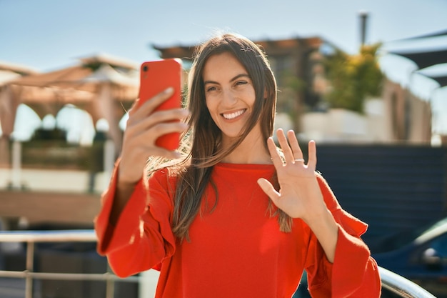 Joven hispana sonriendo feliz haciendo videollamadas usando un teléfono inteligente en la ciudad