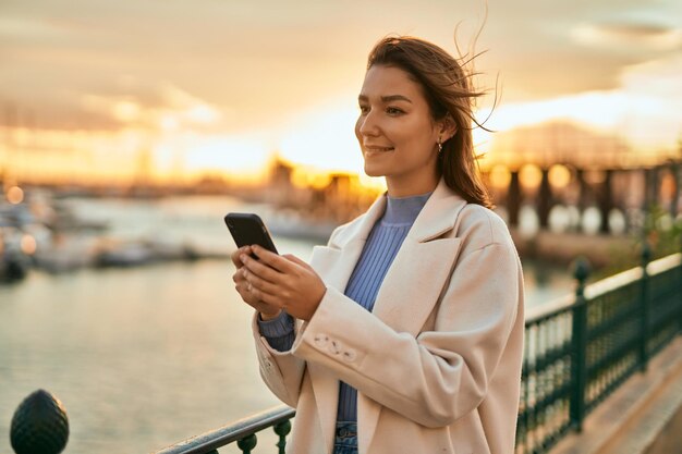 Joven hispana sonriendo feliz usando un teléfono inteligente en la ciudad