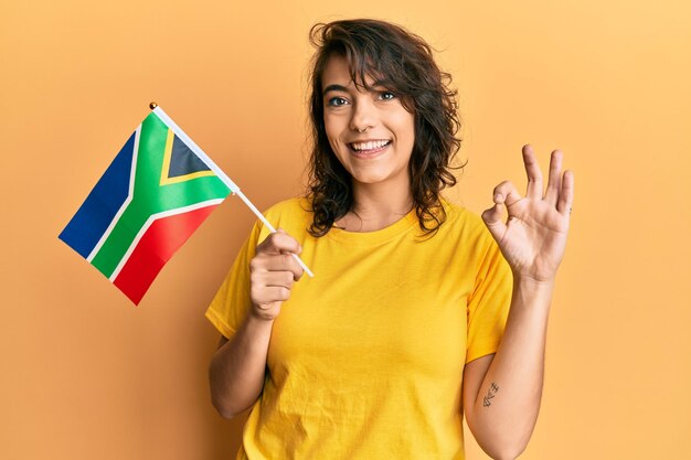 Joven hispana sosteniendo billetes de rand sudafricanos haciendo un buen signo con los dedos, sonriendo amigablemente haciendo un gesto excelente
