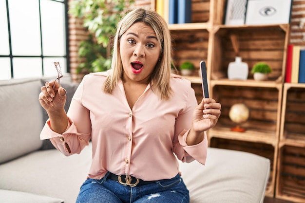 Foto gratuita joven hispana sosteniendo rizador de pestañas y lima de uñas asustada y conmocionada con sorpresa y expresión asombrada miedo y cara emocionada