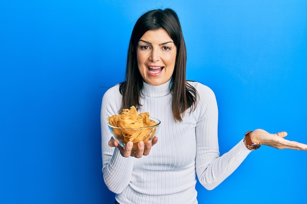 Joven hispana sosteniendo un tazón de pasta sin cocer celebrando el logro con una sonrisa feliz y una expresión ganadora con la mano levantada