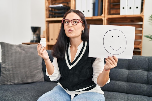 Foto gratuita joven hispana trabajando en la depresión sosteniendo una emoción feliz en papel y pastillas mirando a la cámara lanzando un beso siendo una expresión de amor encantadora y sexy