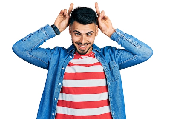 Joven hispano con barba con chaqueta de mezclilla casual posando divertido y loco con los dedos en la cabeza como orejas de conejo, sonriendo alegre