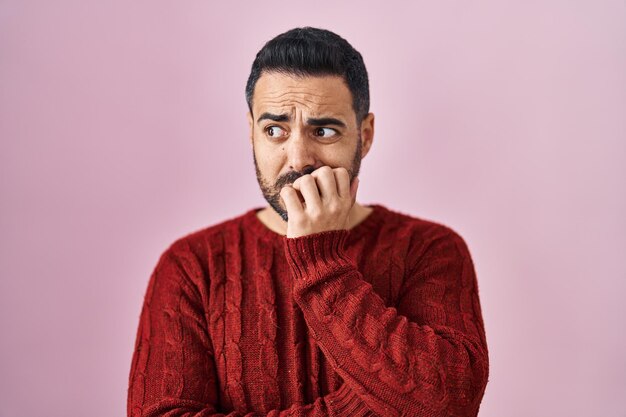Joven hispano con barba y suéter informal sobre fondo rosa que parece estresado y nervioso con las manos en la boca mordiéndose las uñas problema de ansiedad