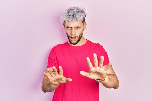 Joven hispano con cabello teñido moderno con camiseta rosa casual haciendo un gesto de parada con las palmas de las manos, expresión de enojo y frustración