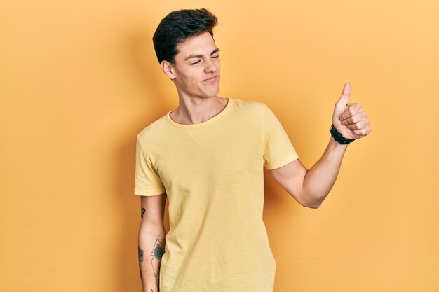 Joven hispano con camiseta amarilla casual que parece orgulloso sonriendo haciendo un gesto de aprobación hacia un lado