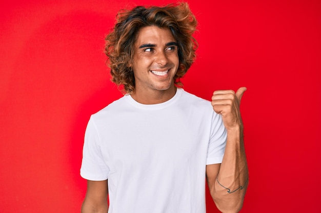 Joven hispano con camiseta blanca informal sonriendo con cara feliz mirando y apuntando hacia un lado con el pulgar hacia arriba.