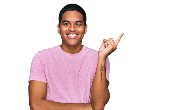 Joven hispano guapo con una camiseta rosa casual sonriendo feliz señalando con la mano y el dedo a un lado