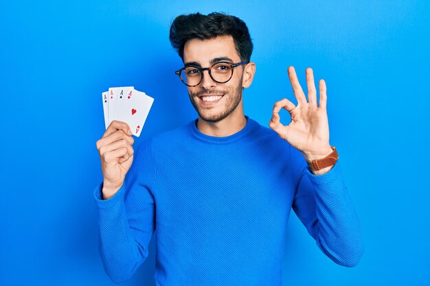 Joven hispano jugando al póquer sosteniendo cartas haciendo el signo correcto con los dedos, sonriendo amable y gesticulando excelente símbolo