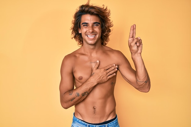 Joven hispano de pie sin camisa sonriendo jurando con la mano en el pecho y los dedos hacia arriba, haciendo un juramento de promesa de lealtad