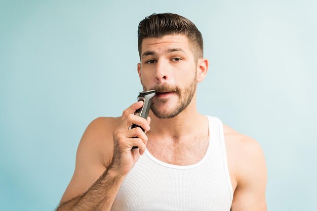 Joven hispano recortando bigote con maquinilla de afeitar eléctrica mientras hace contacto visual con fondo turquesa