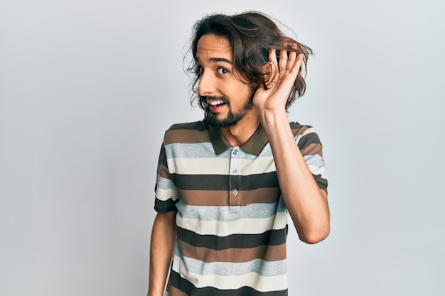 Joven hispano con ropa informal sonriendo con la mano sobre el oído escuchando y escuchando rumores o chismes sobre el concepto de sordera