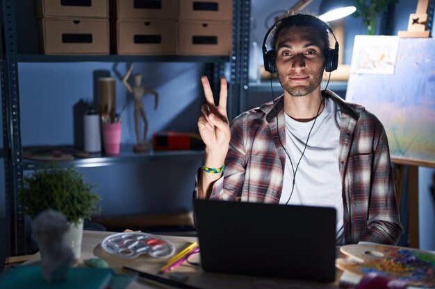 Joven hispano sentado en el estudio de arte con una computadora portátil a altas horas de la noche mostrando y señalando con los dedos número dos mientras sonríe confiado y feliz