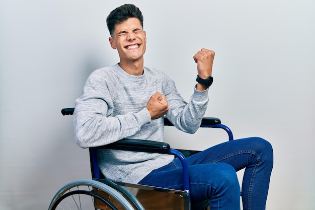 Foto gratuita joven hispano sentado en silla de ruedas celebrando sorprendido y asombrado por el éxito con los brazos levantados y los ojos cerrados concepto ganador