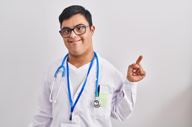 Joven hispano con síndrome de Down vestido con uniforme médico y estetoscopio apuntando con el dedo hacia arriba con una idea exitosa saliendo y feliz número uno