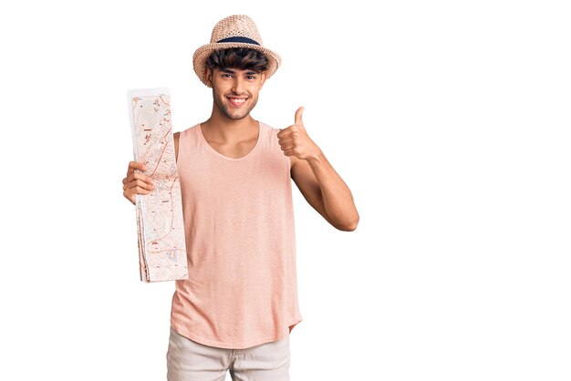 Joven hispano con sombrero de verano sosteniendo un mapa sonriendo feliz y positivo, pulgar arriba haciendo excelente y señal de aprobación
