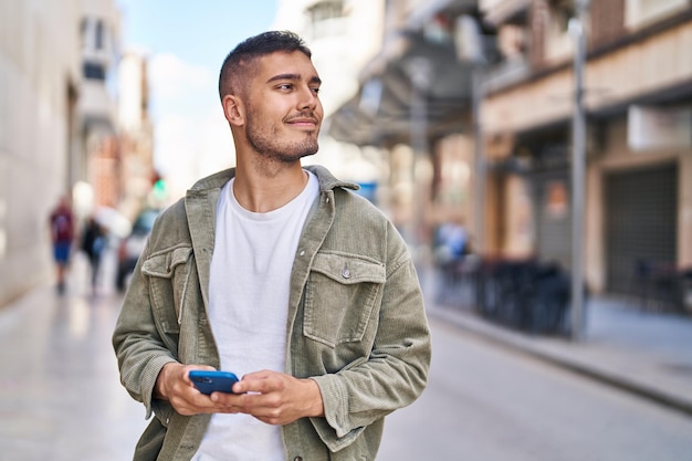 Foto gratuita joven hispano sonriendo confiado usando un teléfono inteligente en la calle