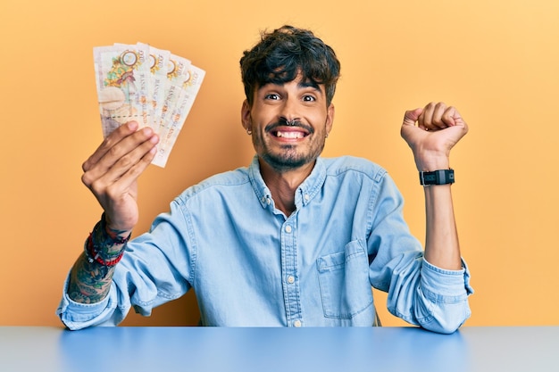 Joven hispano sosteniendo billetes de 10 libras del Reino Unido sentado en la mesa gritando orgulloso celebrando la victoria y el éxito muy emocionado con el brazo levantado