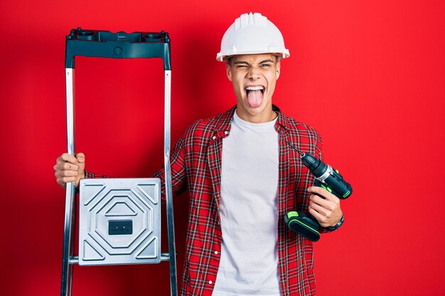 Joven hispano sosteniendo un destornillador usando un casco en las escaleras de construcción sacando la lengua feliz con una expresión graciosa.