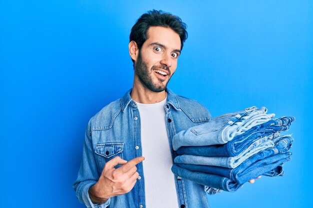 Joven hispano sosteniendo una pila de jeans doblados sonriendo feliz señalando con la mano y el dedo