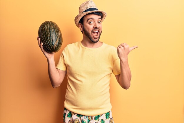 Joven hispano con traje de baño y sombrero de verano sosteniendo melón apuntando con el pulgar hacia el lado sonriendo feliz con la boca abierta