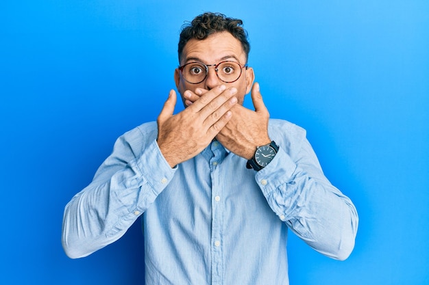 Foto gratuita joven hispano vestido con ropa informal y gafas sorprendido cubriéndose la boca con las manos por error concepto secreto