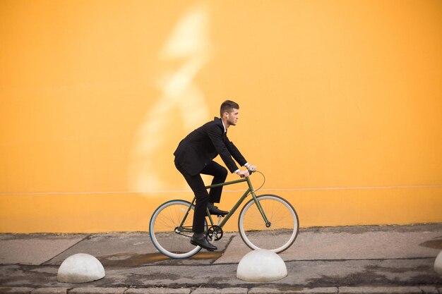 Joven hombre atractivo con traje negro clásico con auriculares inalámbricos montando bicicleta cuidadosamente sobre fondo naranja