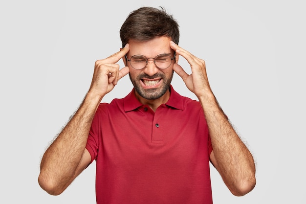 Joven hombre cansado insatisfecho mantiene las manos en las sienes, necesita restaurar la energía después de la noche sin dormir, sufre de dolor de cabeza, está vestido con una camiseta roja, tiene aspecto molesto, aprieta los dientes, se para en el interior