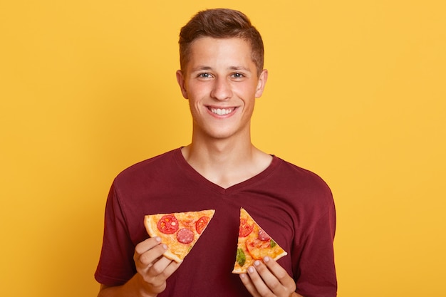 Joven hombre caucásico con dos rebanadas de pizza en el interior, guapo hombre vistiendo casual camiseta marrón posando contra la pared amarilla